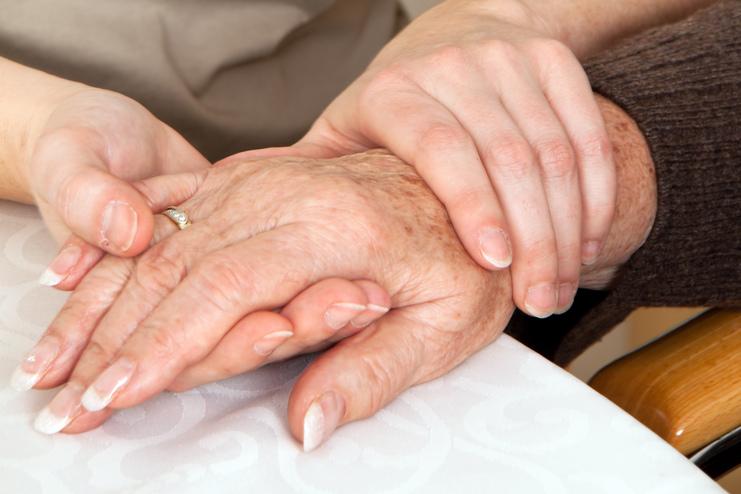 Zwei Hände, eine ältere und eine jüngere, liegen auf einem Tisch, wobei die jüngere Hand die ältere sanft umschließt.